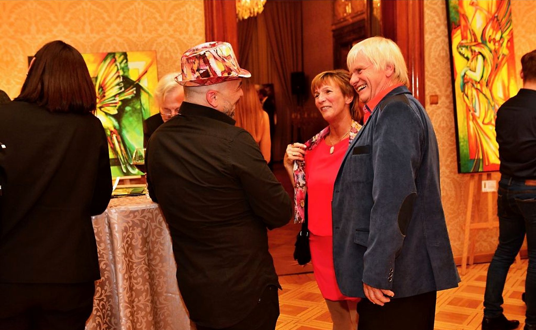 Joe Muczka jr. im Gespräch mit Gästen an einer Vernissage, Werke auf Staffeleien im Hintergrund